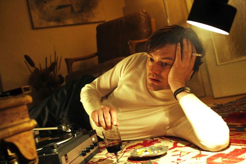 Sam Riley als John Cranko – John Cranko (Sam Riley) liegt daheim neben dem Schallplattenspieler auf dem Fußboden und holt sich, bei Zigaretten und Rotwein, Inspirationen zu neuen Choreografien von immer dem gleichen Musikstück. Was seine wechselnden Lebensgefährten regelmäßig auf die Palme bringt.
