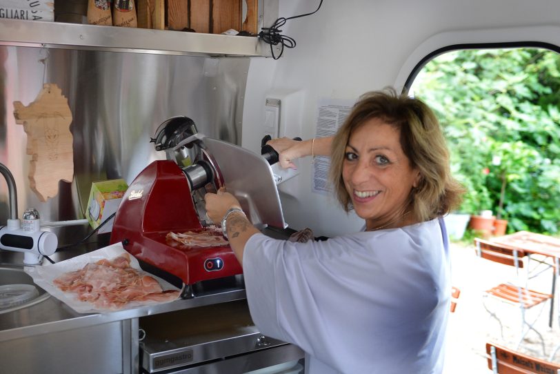 Sardische Leckereien und neue Freundschaften beim ersten Sardischen Abend im Biergarten von Ollis Restaurant. im Bild: Lucia Rosa Cortes von &amp;amp;#039;Sorelle Sarde&amp;amp;#039; schneidet in dem kleinen Wagen, der liebevoll Pitichedda  genannt wird, den sardischen Schinken.