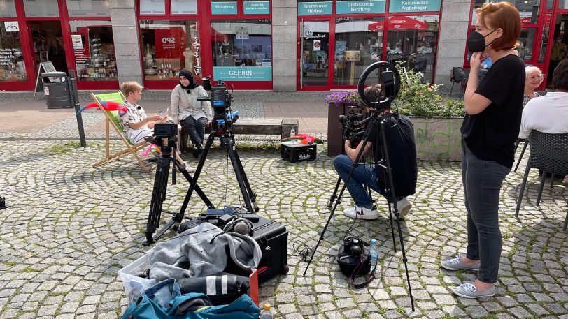 Kamerawinkel und Interverbindung testen: Jan Laser (li.) und Havle Nazik sitzen auf der Bank, Elena Franz (re.) von der Fachstelle Partnerschaft für Demokratie schaut zu.