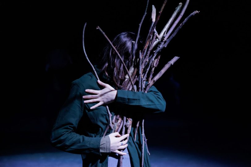 Polina Skarha als Reisigsammlerin, die sisyphosartig aus dem Bündel herausfallende Äste vom Boden wieder aufhebt.