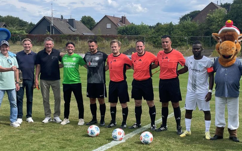 Vor dem Anstoß zum Cranger Kirmes-Cup 2025: Kirmes-Maskottchen Fritz mit seinem neuen Freund Bobby Bolzer, Fußball-Legende Manfred „Manni“ Kaltz (4. von li.), Bezirksbürgermeister Mathias Grunert (2.v.l.), dem Schiedsrichter-Gespann um David Hennig sowie den beiden Kapitänen Collins Agita (SVS) und Toni Curic (Wanne 11).